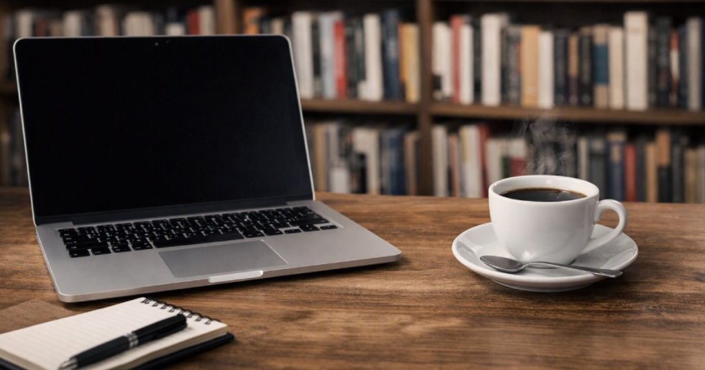 Mesa de madeira com portátil aberto e chávena de café, ao fundo uma estante de livros desfocada.